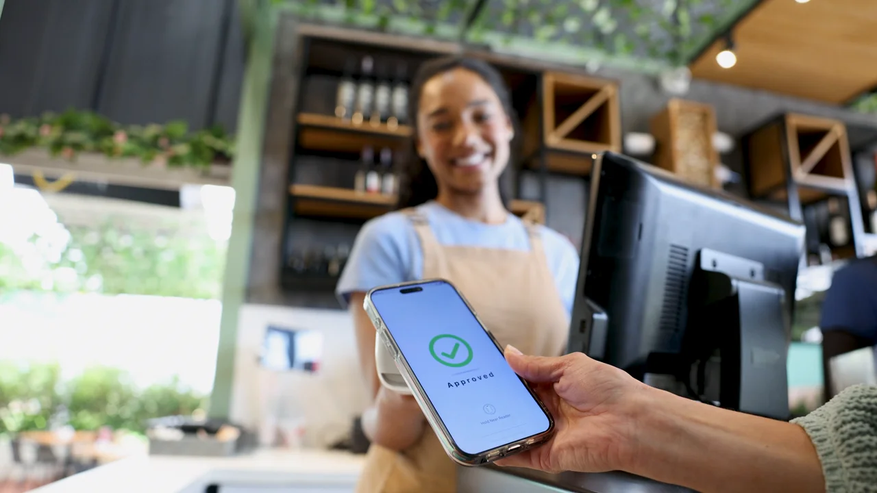 At a cafe counter, a customer holds up a phone showing an approved payment while the server smiles.
